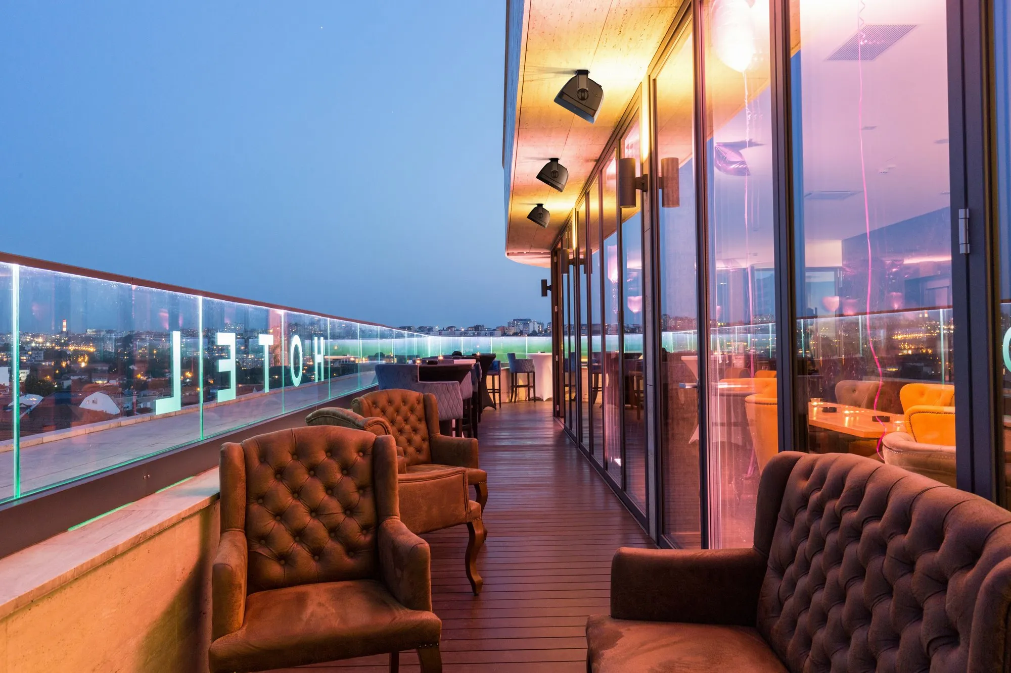 Terrasse de bar sur le toit d'un hôtel au crépuscule, fauteuils capitonnés et enceintes fixées sous l'avancée du toit